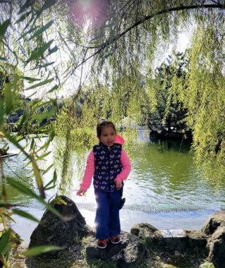 A young girl stands on a rock in front of a lake in Taiwan. She's wearing a long-sleeve pink shirt, a dark vest with hearts on it, and pink shoes. She's surrounded by greenery, with the branch of a tree hanging just behind her head.