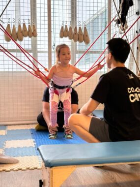 A 5-year-old girl stands on a mat during physical therapy. She's hooked up to a harness that helps her to remain upright while a therapist behind her gently works her ankles. Her father sits cross-legged in front of her. The girl is smiling.