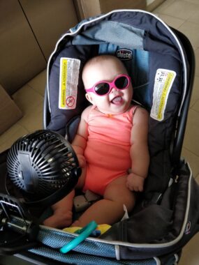 A baby in a pink onesie smiles while seated in a car seat. She's wearing pink-framed sunglasses, and a fan attached to the front of the seat is facing her.