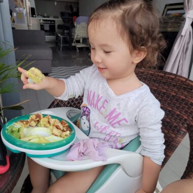 A toddler sits in a high chair, with a meal in front of her on her tray. She has brown hair and wears a white shirt with the words "MAKE ME," in pink, visible. (The other words are obscured.)
