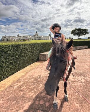 A young girl wearing a black helmet sits atop a horse in a stunning countryside photo in Thailand. It is a sunny day, and the photo is heavily contrasted to show a span of white, puffy clouds in a vast, blue sky. An elegant horse ranch or mansion can be seen in the background. The girl's right arm is raised to the top of her helmet. 