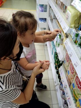 Seen in profile, a toddler picks out an item from a filled store shelf as a woman guides her arms. The brown-haired toddler wears a white T-shirt with a pink shape in the middle. The woman, who seems to be kneeling, wears a black face mask, a black-and-white striped shirt, and black pants. The store's floor is tile in a shade of white.