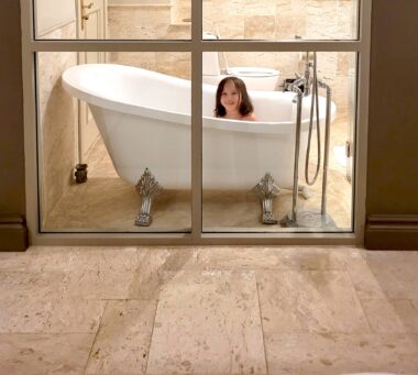 Through four glass panels, we see the head and shoulders of a girl with shoulder-length brown hair in a white bathtub. The surroundings, both in the bathroom and the foreground floors, on the other side of the windows, are tan.