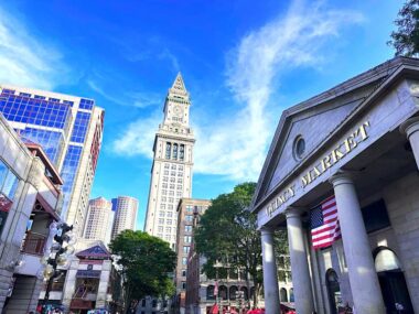 A street-level touristy photo of downtown Boston's Quincy Market. The sun glimmers off the buildings, which stretch to a clear, blue sky. The camera angle accentuates the shapes of the buildings with dramatic angles, lines, edges, and rooftops. 