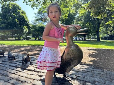 A young girl in a pink blouse and pink and white dress poses with a giant duck statue (with other ducks lined up behind). It's a "Make Way for Ducklings" statue in Boston, in what appears to be a park. Several shade trees allow the bright sun to cast broad shadows, giving the sense of both summertime fun and the relaxation of a shaded park. 