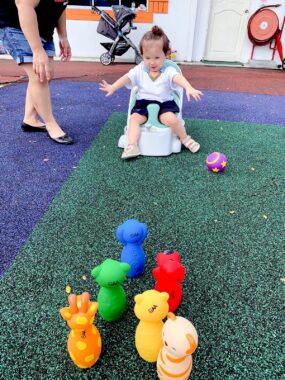 At the end of a rectangle of green artificial grass, we see a toddler in a white top and black shorts sitting on what appears to be a small white chair, with an aqua-colored extension between her upper legs. We see the legs, arms, and midsection of a woman in jean shorts and a black top at right, while toys in various colors are in the foreground. One violet ball is near the toddler's feet. The flooring area surrounding the turf is purple, with dark pink just beyond it. The walls are white.
