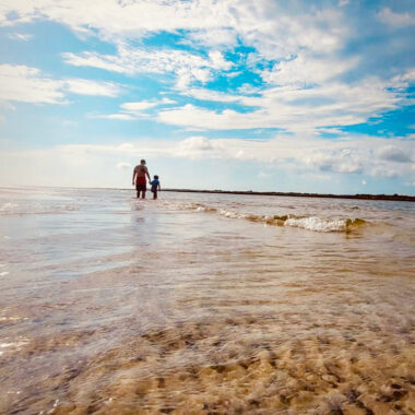 A sharp image that could be on a travel brochure depicts a father and daughter off in the distance, walking in ankle-deep water on the beach. The clear water and sandy beach stretch to the horizon, where they blend into a blue sky with fluffy white clouds. The camera's low-to-the-ground angle makes for a stunning shot that stretches the scenery seemingly to infinity. The tiny image of a man and his daughter in the distance is also quite touching. 