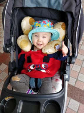 An infant sits in an all-black stroller. She is smiling and wears a green, wide-brimmed hat with shapes in different colors. Tan and brown cushions are on either side of her head. A red monster clothing is on her torso, appearing to be over a navy blue T-shirt. She wears gray pants. The stroller is sitting on a pink, off-white, and brown tile floor.