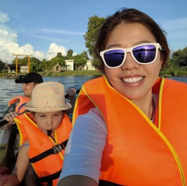 On a lake with greenery and a house in the background and blue sky overhead, three people ride in a boat. The woman, in front, has dark hair and wears purple-tinted sunglasses; the child in the center has on a straw hat; and the man in back wears a black baseball cap. All three wear orange life vests.
