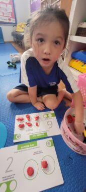 A child sits on blue floor mat and faces the camera, wearing navy blue shorts and a navy blue T-shirt with white on the shoulders and the top of the sleeves. One of her hands is reaching into a pink container, and in front of her are two white cards with some green units; circles of the cards are topped with what appears to be red dough. 