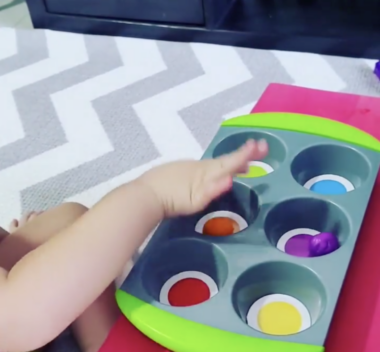 A child's hand reaches for what looks like a gray metal muffin tray with six spots, each with a different color at the bottom. It appears to be on a pink platform, with a surface of gray and white stripes in front of it.