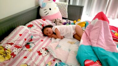 A 5-year-old girl curls up in her bed atop pink striped Hello Kitty sheets. She's lying on her side, with her hands folded under her head. A comforter partially covers her legs, and there's a large stuffed Hello Kitty toy sitting up in the bed beside her. It appears to be midday, as sunlight is streaming in under the curtains.