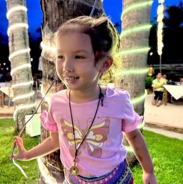 A little girl wears a pink shirt with a brown, yellow, and white butterfly on it as she stands in a grassy area with trees decorated in rings of light. She wears a black cord around her neck with a pendant hanging down near her navel. In the background is what appears to be a tile area with tables, with an evening blue sky and trees beyond it.