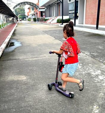 A young girl on a purple scooter travels down a concrete outdoor alley of sorts, her back to the camera. She wears a bright red shirt with loose, multicolored short sleeves and what appears to be a blue jean skirt or shorts. An arch is in the distance.