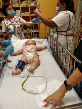 A baby in a pink shirt and diapers lies on a cot with tubes attached to her nose and a monitor on her leg. At the head of the bed are two hospital workers; a pair of blue-gloved hands reach out to her on the left. 