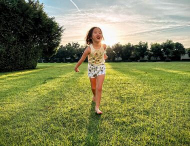 A young girl runs across a large, grassy field toward the camera. The morning sun is directly behind her, creating a glow around her head. She's barefoot and wearing a yellow tank top and blue and white shorts.
