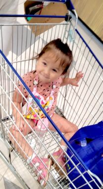 A young girl sits inside a shopping cart next to what appears to be a blue backpack or shopping bag. She's wearing a pink outfit and grinning at the camera.