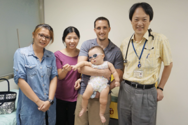 Two women and two men stand facing the camera in a room with a pale tan wall behind them. From left are a woman in either a blue skirt or long shirt; a woman in a purple shirt and black pants; a man in a gray shirt and khakis, who's holding a baby who wears a white onesie and sunglasses; and a taller man in a yellow shirt and gray slacks, with a lanyard and what appears to be a stethoscope around his neck.