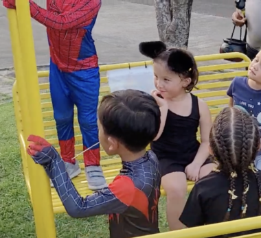 A young girl wearing a black leotard and black cat ears sits on a yellow park bench, smiling at someone off to her right. She's surrounded by several other children wearing Halloween costumes.