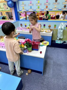 A young girl stands across from a boy about her age at an indoor play center. There's a low counter between them covered in various play objects, and the children seem to be having a conversation.
