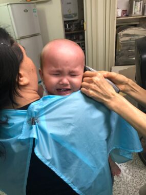 A baby has her head shaved in a hospital setting while being held by her mother. 