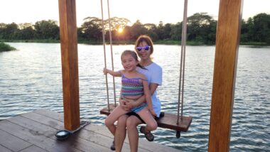 A young girl sits on her mother's lap on a swing next to a body of water, such as a lagoon or river. The sun appears to be setting in the background. 