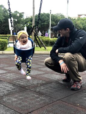 A father crouches down to supervise a toddler in a swing for babies at an outdoor park. The young girl has her mouth wide open in happiness and awe. 