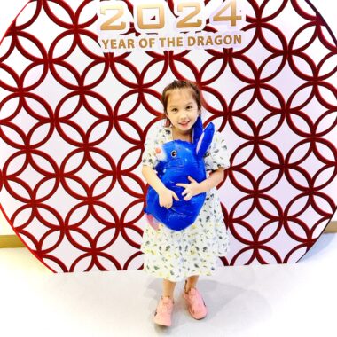 A young girl wearing a dress poses for a photo while hugging a blue stuffed animal. She's standing in front of a circular display that reads "2024: Year of the Dragon."