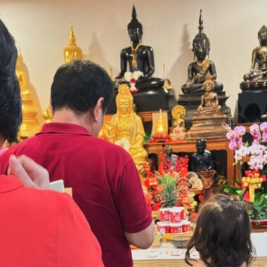 A young girl stands next to her grandparents in a temple to pray and receive blessings on the Lunar New Year. The photo is taken from behind, and in front of the three people pictured are a variety of Buddha statues, most of which are black or gold.