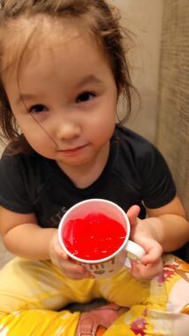 A young girl in a black top and yellow pants holds a small mug that appears to be full of red juice. 