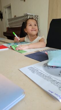 A young girl sits at a table and completes her schoolwork. She's holding a green marker and has a workbook open in front of her, with other school supplies spread out around her. She's smiling up at someone off camera.