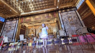 A low-angle photo shows a young girl standing in the middle of a large, empty room and singing. The space appears to be some sort of temple, with chairs spaced evenly around her, and a platform with statues in the background.