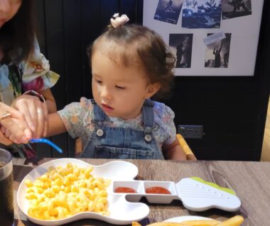 A young girl sits in a high chair as her mother, just out of frame, helps her grasp a blue utensil. They're seated at a table, and in front of the girl is macaroni and cheese in a bowl shaped like a guitar.