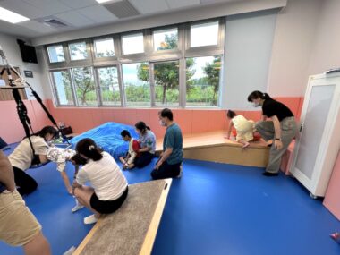 A room holds about 10 people – some adults and some children – who are engaged in various activities. There is a wooden ramp, a large swing, and other equipment for the children to use.
