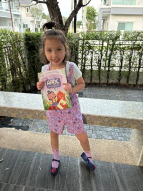 A young girl in purple overalls stands beside a stone bench in front of a small courtyard. She's holding up a children's book about AADC deficiency and smiling.