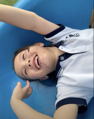 A young girl with a white top and blue collar is seen from above on a blue surface. She is grinning and has outstretched arms.