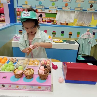 A young girl with dark hair wears a green and white cap, a pink blouse, and a pale-green overshirt while handling a pretend ice cream cone. She's in a colorful play space that looks designed to be an ice cream parlor.