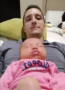 A man lies on a bed with his infant daughter sleeping on his chest. She's wearing a pink onesie while her dad is in a striped polo shirt. He's resting his head on a yellow-green pillow and taking the photo from a low angle.