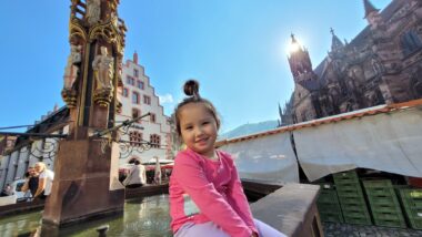 A young girl in a pink shirt smiles at the camera while sitting on a ledge next to a fountain in German town. The wide-angle photo captures many striking buildings in the background