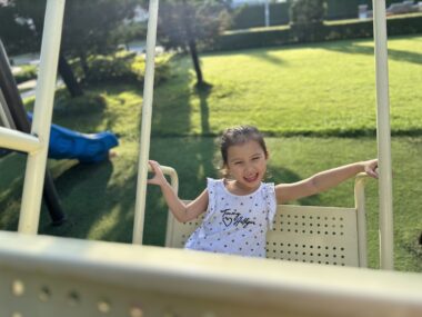A young girl laughs while seated on a large metal swing. The angle of the photo suggests that her father is pushing her. There's a sunlit lawn in the background, and a blue slide is visible on the left.