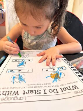 A young girl, maybe 3 or 4 years old, sits at a table and colors in a worksheet. The assignment shows pictures of six objects and asks the child to select which letter the object starts with. The girl has circled several letters and colored in a few of the objects, and appears very focused on her work.