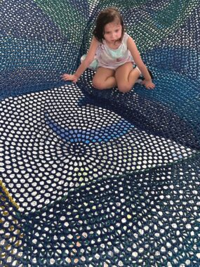 A young girl kneels on a large blue net at the playground. She's frowning and not making eye contact with her dad, who's taking the photo.