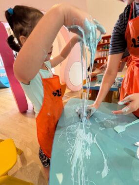 A young girl in an orange apron stands in front of a table while playing with turquoise-colored slime. There's a glob of it on the table, and she's pulling up long strands of it, her hands covered in slime and at the height of her head.