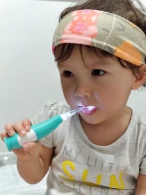 A young girl is putting a green and white toothbrush into her mouth. She wear a gray T-shirt with writing on it and largely green bandana.