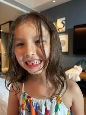 A young girl with longish brown hair grins at the camera, revealing an empty space in the center of her bottom teeth. She's wearing a white outfit with straps and multicolored images.