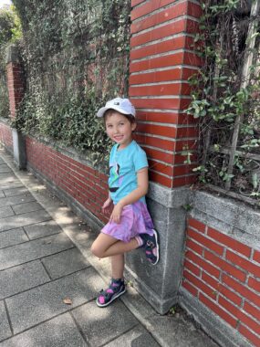 A young girl leans against a brick wall with her left foot up against the wall. She appears to be in an outdoor area like a large garden.