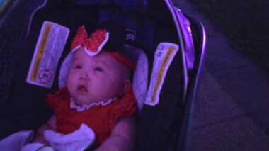 A newborn sitting in a baby stroller stares up at something in the distance. She's wearing all red, including a red bow on her forehead.