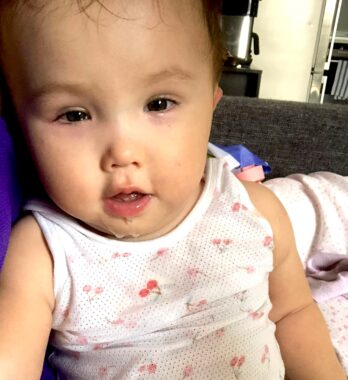 A baby looks at the camera in a close-up photo. She's wearing a white tank top with a cherry pattern and has drool running down her chin.