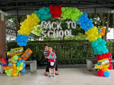 A young girl stands in front of her mom under a large, colorful arch of balloons. Just under the top of the arch are letter balloons that read "back to school." The mom is crouched down so her face is next to her daughter's, and both are smiling broadly.