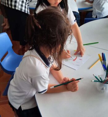 A first grader sits at a round table holding a pencil. She's watching a girl to her left, who is coloring on a sheet of paper. Both are in white and navy school uniforms.